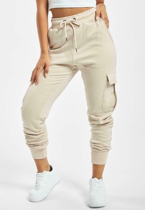 Mujer con joggers cargo beige con cintura ajustable con cordón y zapatillas blancas, de pie con una mano en el bolsillo.