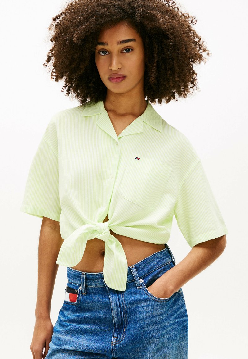 Ung kvinde med krøllet hår iført en lysegrøn kortærmet skjorte, der er slået op i taljen, og blå højttaljede jeans.