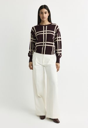 Woman wearing a dark sweater with beige grid pattern and white wide-leg trousers standing against a plain light background.