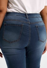 Blå denimjeans med en figurnära design, med bakfickor och kontraststickning. Tyget har ett slätt, något texturerat utseende.