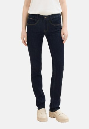 Vaqueros de mezclilla azul oscuro con un corte slim, que cuentan con cinco bolsillos, costuras en contraste y una textura suave. Llevados con zapatos beige.
