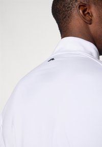 Weißes Sportshirt mit hohem Kragen, ausgestattet mit einer glatten Textur und einem kleinen schwarzen Logo auf der Rückseite in der Nähe des Kragens.