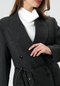 Donkergrijze visgraat dubbele rij knopen jas met zwarte knopen en een zwarte ceintuur in de taille. Gecombineerd met een witte top met hoge hals. Gladde textuur.