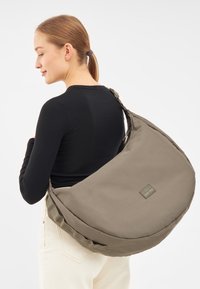 Borsa a tracolla in morbido tessuto marrone, design curvo con un ampio scomparto, tracolla regolabile e una patch rettangolare con logo sul lato.