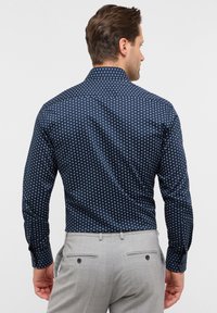 Camicia blu navy con un piccolo motivo geometrico bianco, dotata di colletto classico e maniche lunghe. Indossata con pantaloni grigi chiari.