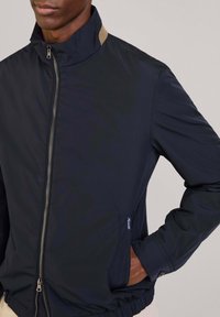 Chaqueta cortavientos de color navy con un cuello alto, cremallera frontal, puños elásticos y un bolsillo lateral con un acento marrón. Tejido elegante y resistente al agua.
