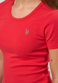 Lähikuva henkilöstä, joka pukeutunut istuvaan, punertavaan, ribbineulottuun lyhythihaiseen paitaan, jossa on pieni kirjailtu logo rintakehässä: hevosen selässä oleva polo-pelaaja.