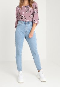 Blus med blommönster i mauve färg och trekvartslånga ärmar, kombinerad med ljusblå högmidjade jeans och vita sneakers.
