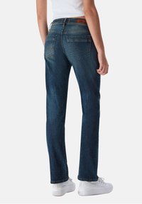 Jeans de mezclilla azul oscuro con corte recto, bolsillos traseros detallados y áreas ligeramente desgastadas. Combinados con zapatillas blancas.
