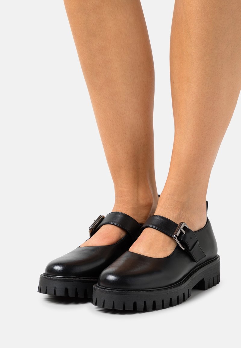 Chaussures Mary Jane en cuir noir avec semelles épaisses crantées et brides réglables à boucle métallique, portées pieds nus.