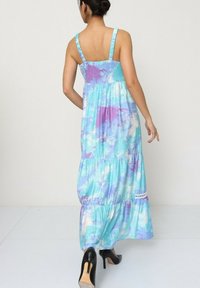 Robe maxi avec un corsage smocké, des bretelles ajustables et une jupe fluide à volants en bleu avec des motifs aquarelle violets. Portée avec des talons noirs.