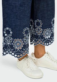 Donkerdenim wijde broek met witte bloememblemen en ronde uitsparingen langs de zoom, gecombineerd met witte sneakers.