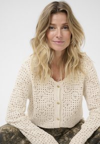 Blonde Frau mit welligem Haar, die eine cremefarbene Häkeljacke und gemusterte braune Hosen trägt, sitzt und schaut in die Kamera.