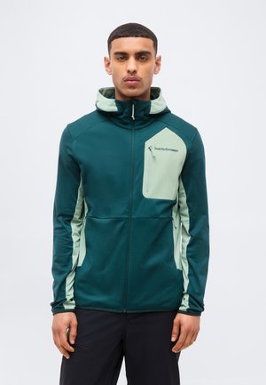 Mees, kes kannab türkiissinist ja heledat rohelise lukuga kapuutsiga jopet rinnataskuga ja "Peak Performance" logoga, seisab tavalise tausta ees.