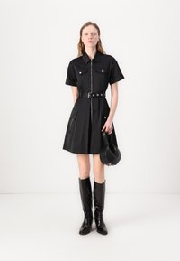 Vestido preto de manga curta com fecho de correr à frente, dois bolsos no peito, cintura cintuada e saia rodada. Combinado com botas pretas brilhantes até aos joelhos.