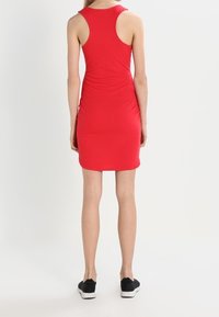 Robe rouge ajustée avec un design sans manches et un col dos nageur. Tissu lisse et extensible avec des fronces à l'arrière. Portée avec des baskets noires.