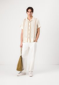 Cămașă crem cu mânecă scurtă, având un model texturat în nuanțe de bej deschis, combinată cu pantaloni albi și o geantă de umăr kaki. Tenisii albi completează ținuta.