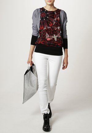 Top met lange mouwen in meerdere kleuren met bloemen- en vlinderpatronen, gecombineerd met witte skinny broek en een zilveren, gestructureerde handtas.