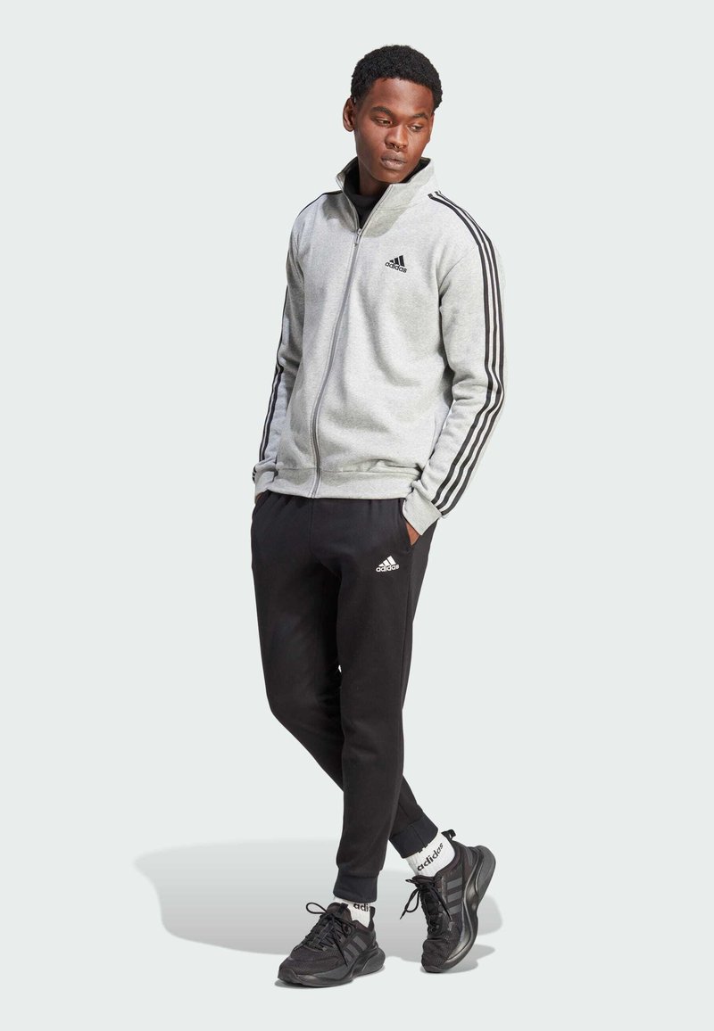 Chaqueta Adidas gris con cremallera y rayas negras, combinada con joggers negros que tienen un pequeño logo y zapatillas deportivas negras con detalles blancos.