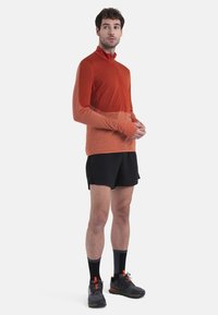 Maglietta sportiva a maniche lunghe con zip in gradiente arancio, dotata di fori per i pollici, abbinata a pantaloni corti neri e sneakers nere con vivaci dettagli arancioni.