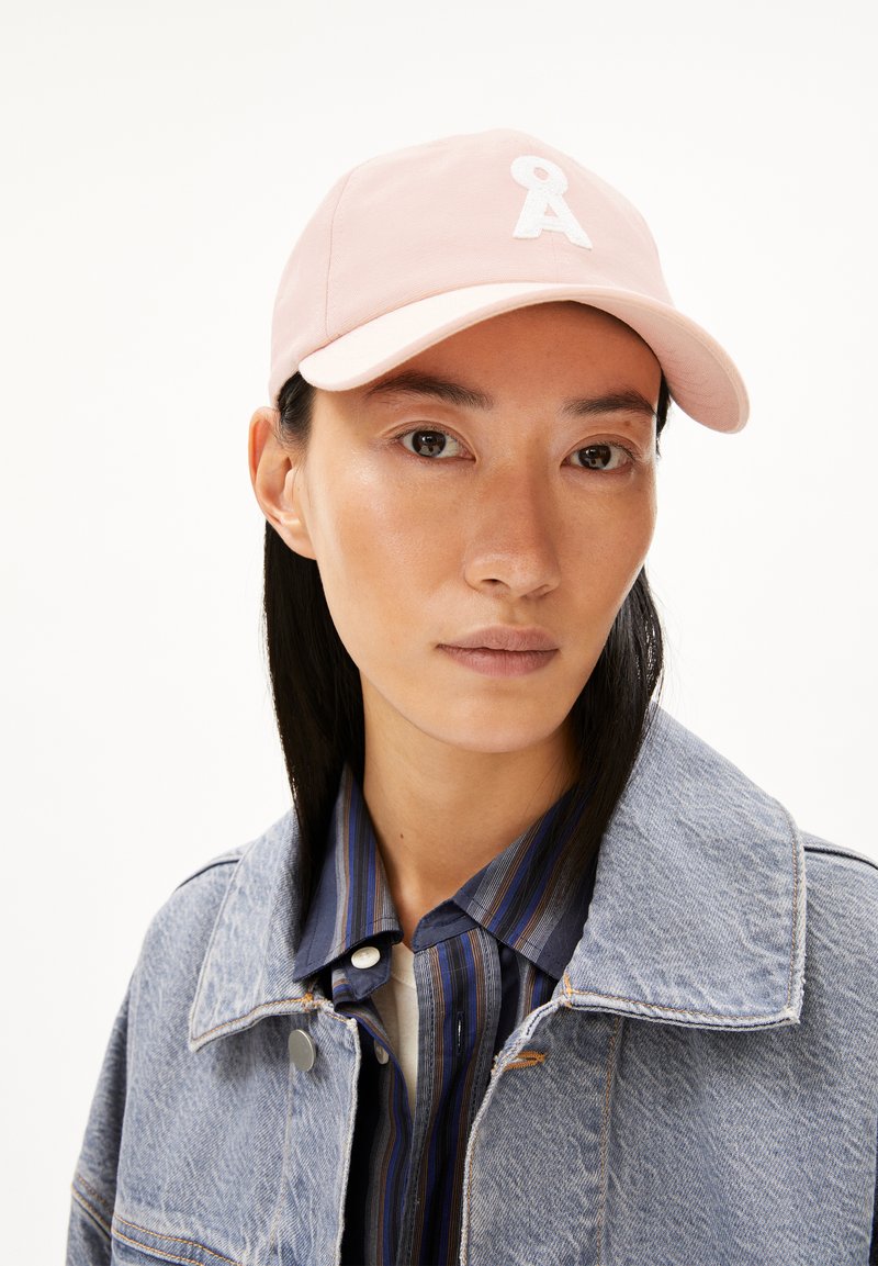 Lichtroze baseballpet met een geborduurde witte letter "A". Van katoen, met een gebogen rand, gedragen met een denimjack en een gestreept shirt.