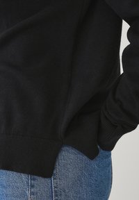 Maglione nero a maglia con vestibilità comoda e polsini a righe, abbinato a jeans blu chiaro per un look informale. Il maglione presenta una consistenza morbida.