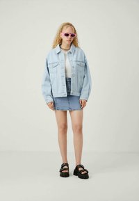 Casaco de ganga azul claro, top branco, saia de ganga azul, sandálias de plataforma pretas, óculos de sol cor-de-rosa, colar de corrente prateado; outfit descontraído em camadas.