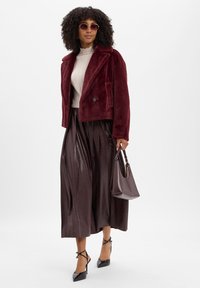 Vrouw met een bordeauxrode nepbontjas, een crèmekleurige geribde top, een geplooide donkerbruine rok, een zonnebril, zwarte hakken en een bordeauxrode handtas.