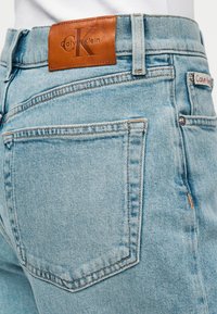 Ανοιχτό μπλε τζιν με καφέ δερμάτινο λογότυπο Calvin Klein στη μέση. Διαθέτει τσέπες και ραφές στη βασική σχεδίαση.