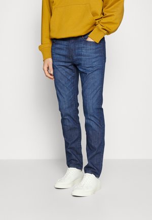 Person iført blå slim-fit jeans, hvide sneakers og en sennepsgul hættetrøje med den ene hånd i lommen.