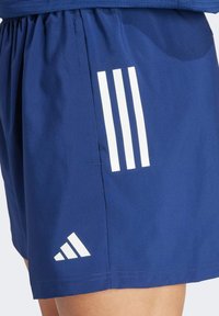 Námornícke modré športové šortky vyrobené z hladkej tkaniny; obsahujú tri vertikálne biele pruhy a biely logo trojuholník blízko lemu.