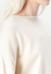 Beige breipullover met een ribbelrand bij de hals en een zachte textuur. Het kledingstuk heeft een comfortabele pasvorm en verlaagde schoudernaden.
