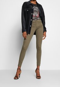 Camisa chaqueta de pana negra sobre una camiseta gráfica, jeans skinnies verde oliva y tacones altos negros con tiras. Textura suave, diseño ajustado.