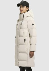 Lunga giacca trapuntata beige con collo alto, tasche con zip, coulisse regolabile e patch rotondo con logo sulla manica.