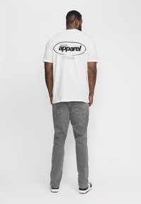 Camiseta blanca de manga corta con un gráfico negro en la parte posterior, combinada con jeans grises y zapatillas en blanco y negro. Fondo liso.
