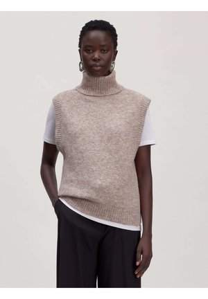 Vrouw draagt beige gebreide mouwloze coltrui-vest over wit kortermig shirt en zwarte broek, staand tegen een effen achtergrond.
