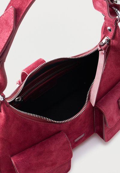 Τσάντα από κόκκινο σουέντ με ασημένιο φερμουάρ, που διαθέτει έναν ανοιχτό κύριο χώρο και δύο μπροστινές τσέπες με καπάκια σε λευκό φόντο.