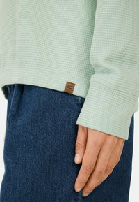 Lichtgroene, textuurrijke longsleeve shirt met een geribbeld patroon, gecombineerd met donkerdenim jeans. Heeft een bruine label nabij de onderzoom.