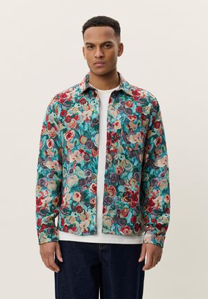 Bărbat purtând o cămașă colorată cu model floral peste un tricou alb și blugi închiși la culoare, stând în picioare pe un fundal simplu.
