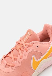 Vaaleanpunaiset urheilukengät, joissa on verkkokudosta ja teksturoituja materiaaleja, keltaisella swoosh-logolla, monimutkaisella nauhoitusmuotoilulla ja pehmustetulla nilkalla.