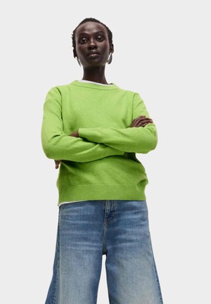 Pull en maille verte à col rond, avec poignets et ourlet côtelés. Porté sur un t-shirt blanc, associé à un jean coupe large bleu clair.