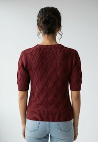 Bordowy sweter z krótkim rękawem z teksturowanym wzorem warkocza oraz prążkowanym dekoltem i dołem, zestawiony z jasnoniebieskimi dżinsami.