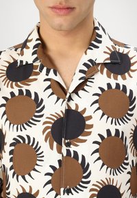 Camicia a maniche corte in tessuto crema con grandi motivi a raggiera marroni e neri. Include un colletto a punta e bottoni neri.