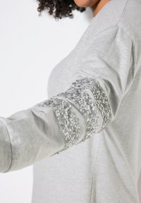 Felpa grigia con maniche lunghe e paillettes silver texturizzate lungo il braccio, creando un contrasto sorprendente con il tessuto morbido.