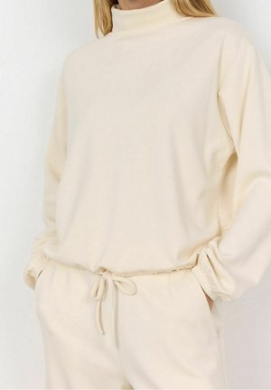 Mujer con sudadera de cuello alto color crema y pantalones a juego con cordón, con las manos en los bolsillos, frente a un fondo liso.