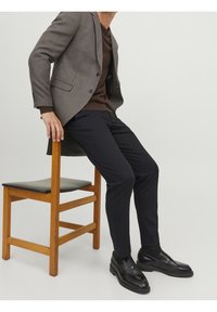 Grau kariertes Blazer über einem braunen Pullover, kombiniert mit schwarzen Hosen und Mokassins. Ein Stuhl aus Holz mit einer schwarzen Sitzfläche befindet sich im Vordergrund.