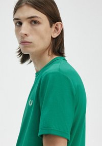 T-shirt in cotone verde con colletto tondo e maniche corte, caratterizzato da un piccolo logo ricamato sul petto. La texture appare liscia.