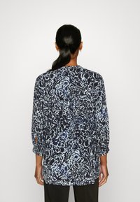 Mujer con coleta negra que lleva una blusa de manga larga suelta de color azul marino y azul claro con un diseño, de espaldas contra un fondo blanco.