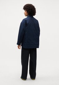 Giacca oversized di colore blu navy con collo, caratterizzata da una texture liscia e due bottoni laterali, abbinata a pantaloni scuri a gamba larga.