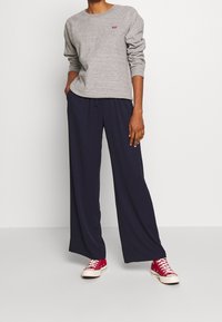 Felpa grigia con collo rotondo e logo rosso; pantaloni larghi blu navy; sneakers di tela rosse. Un outfit semplice e casual con colori a contrasto.